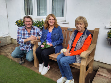 teak garden bench with 2 ladies seated on it on a patio with grass lawn