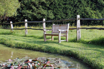 outdoor garden bench on grass with a fence in the background overlooking a pond