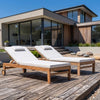 Two wooden outdoor sun loungers with wheels and white cushions are arranged on a weathered wooden deck in front of a modern house with large windows and wood paneling.