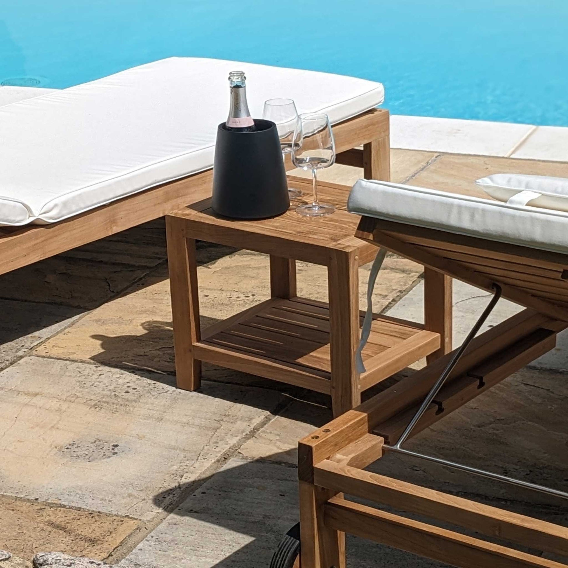 Wooden teak garden side table, shelf with wine glasses and a bottle on a patio by a poolside.