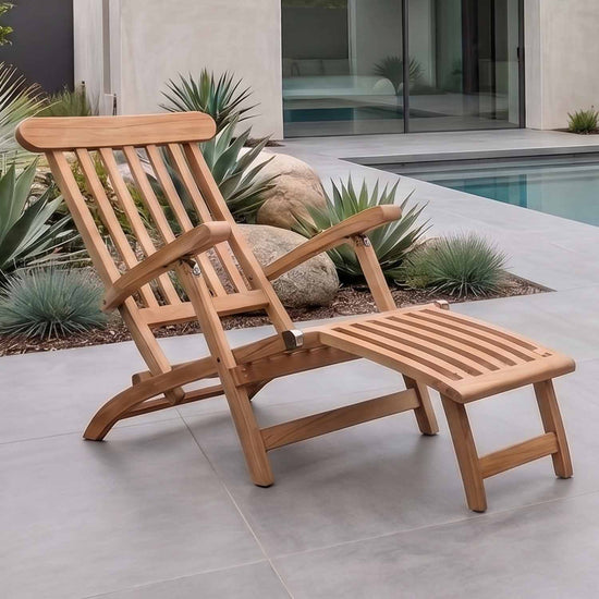 A wooden outdoor teak steamer chair on a tiled patio beside a pool with plants and rocks in the background.