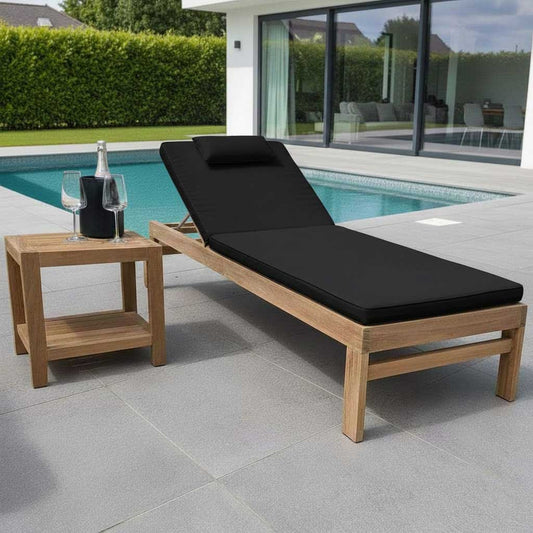 Wooden sun lounger with black cushion next to a wooden side table by a poolside.