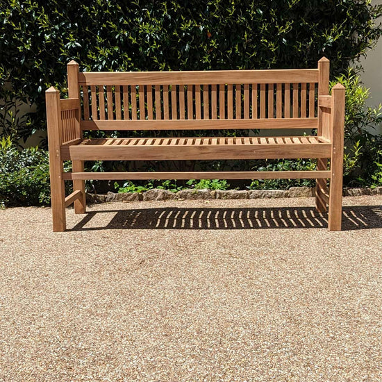 wooden teak garden bench, contemporary architectural design, on a gravel surface with greenery in the background