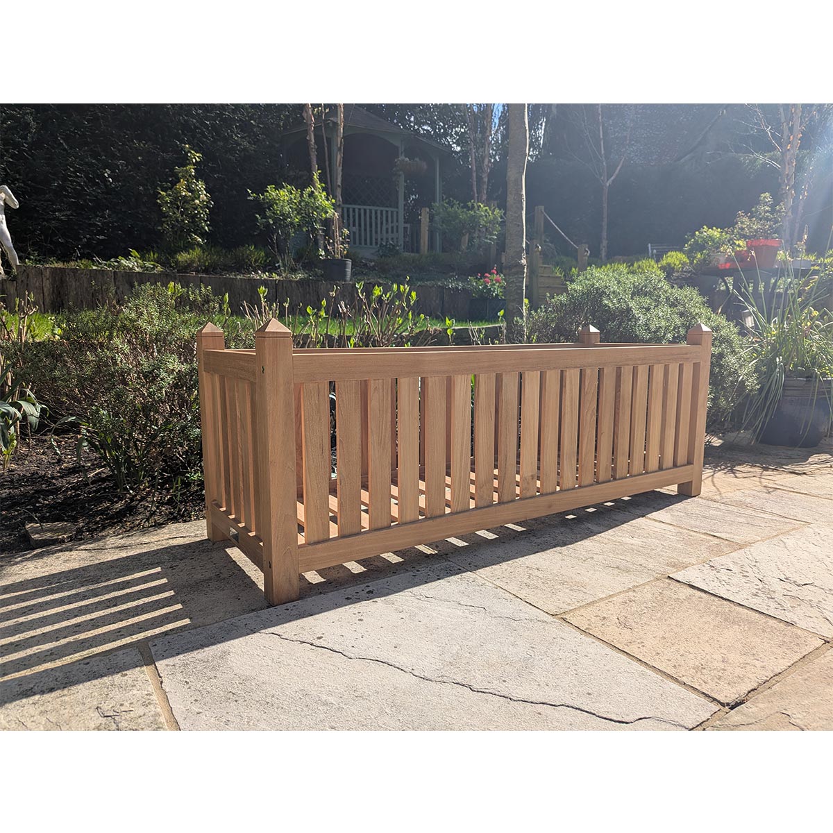 Wooden teak planter box on a patio with garden and trees in the background