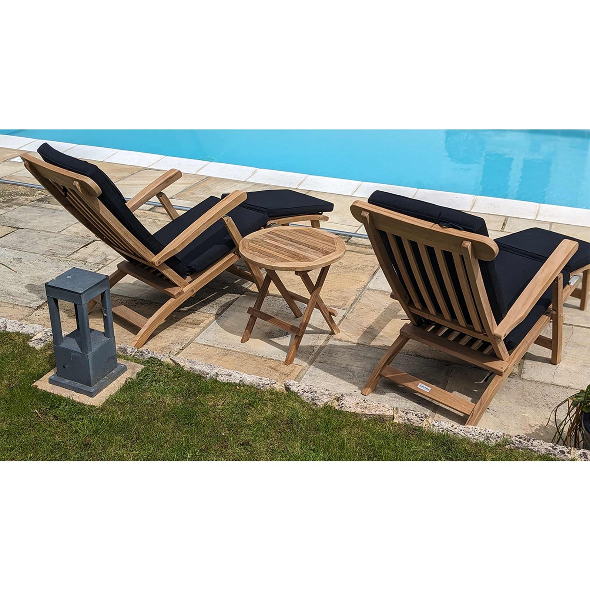 Two wooden lounge chairs with black cushions and a round folding coffee table by a poolside.