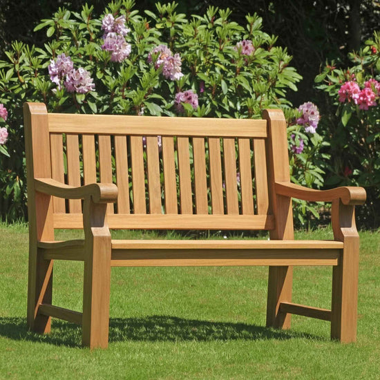Wooden 2 seat teak garden Great Maytham bench on grass with flowering bushes in the background