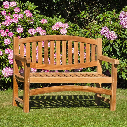 Wooden oval 2 seat garden bench, Marden, in a garden setting with pink flowers and greenery