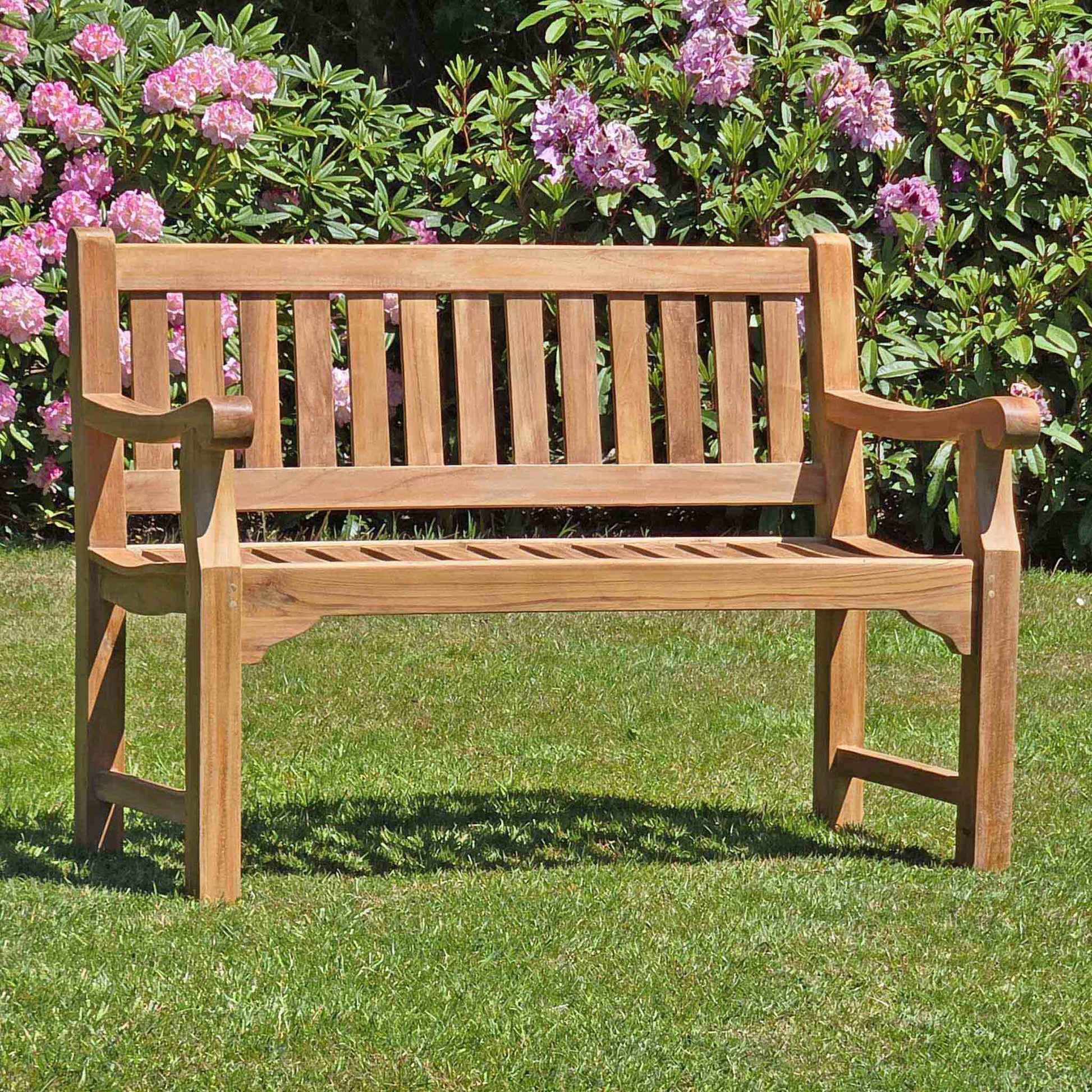 classic 2 seat teak garden bench, Sandhurst, in a garden setting with pink flowers and greenery