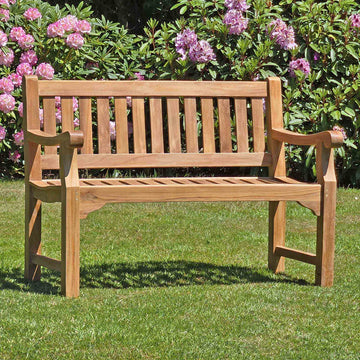 classic 2 seat teak garden bench, Sandhurst, in a garden setting with pink flowers and greenery