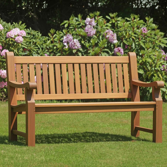 Heavy duty teak wooden bench in a garden with green grass and purple flowers in the background