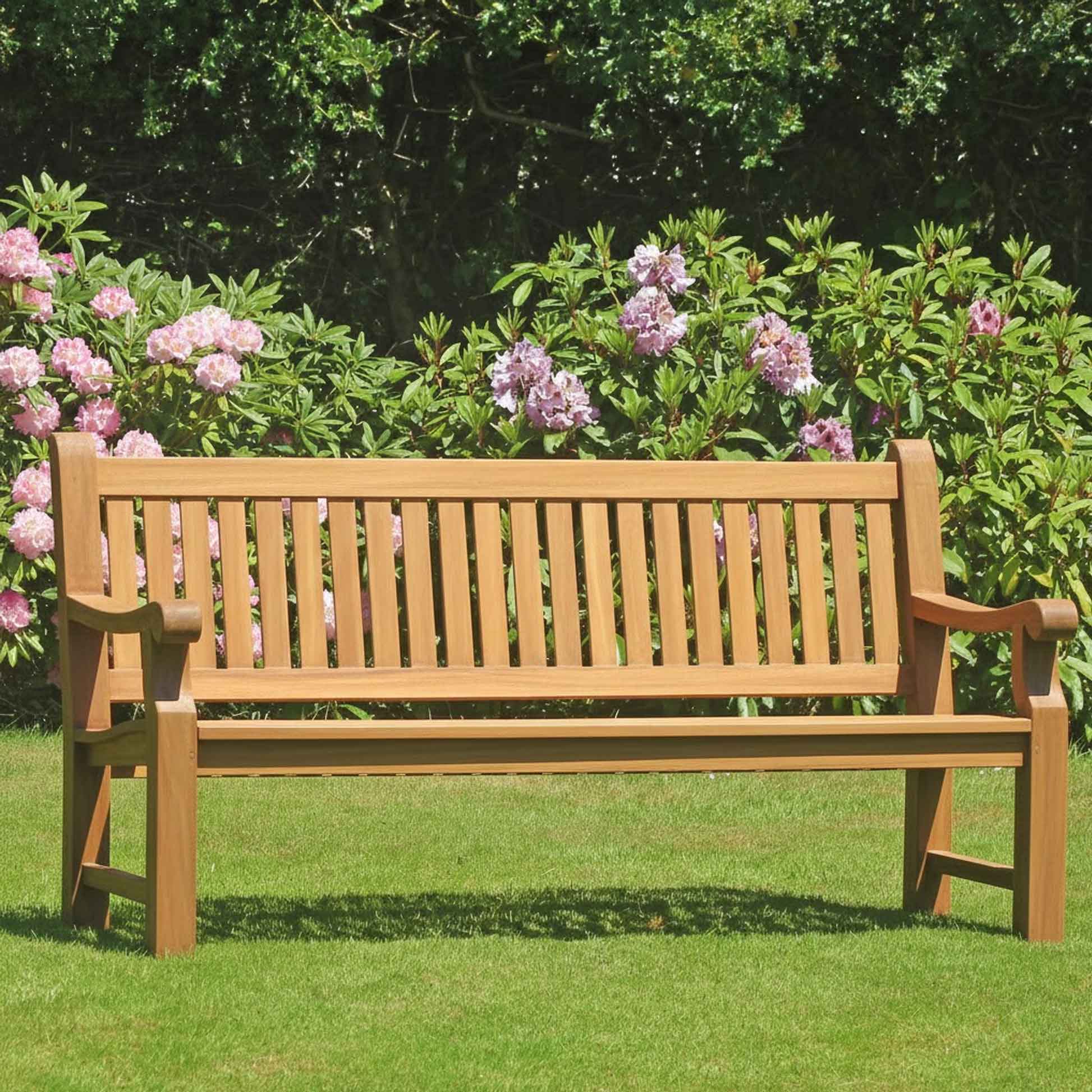 Wooden 4 seat teak garden Great Maytham  bench in a garden setting with purple rhododendrons in the background 