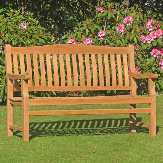 Wooden teak garden bench, 3 seat Scotney, with a high arched back in a garden setting with green grass and pink flowers.