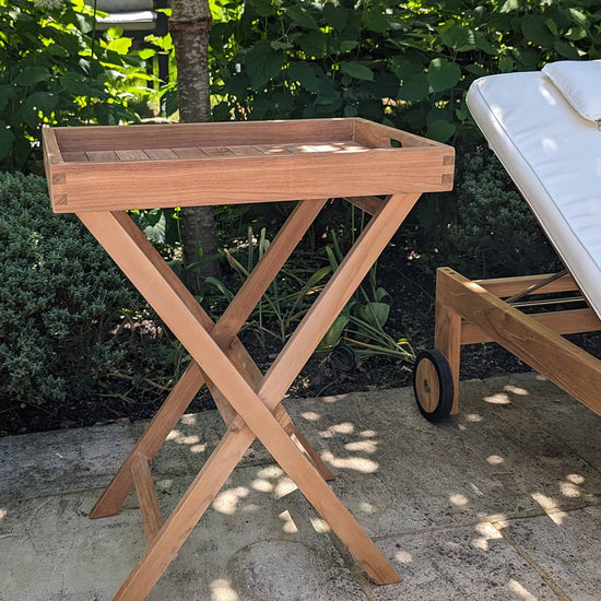 Wooden serving tray on a stand in an outdoor setting with a lounge chair and greenery.