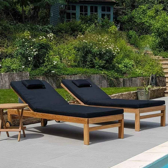 pair of teak garden sun loungers with black cushions on grey ceramic tiles and by a pool