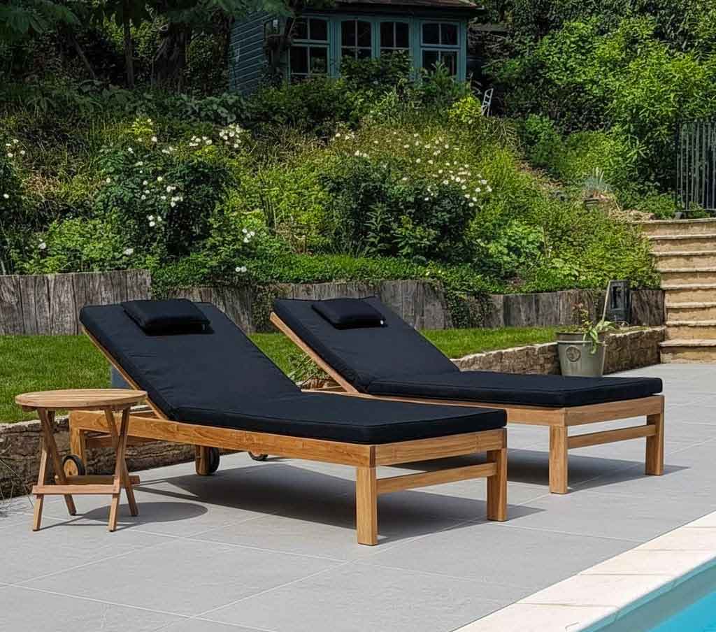 pair of teak garden sun loungers with black cushions on grey ceramic tiles and by a pool