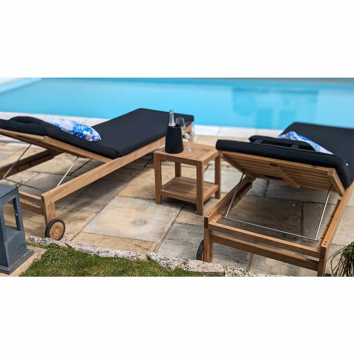 Pair teak wood sun loungers, with wheels,  black cushions by a poolside 